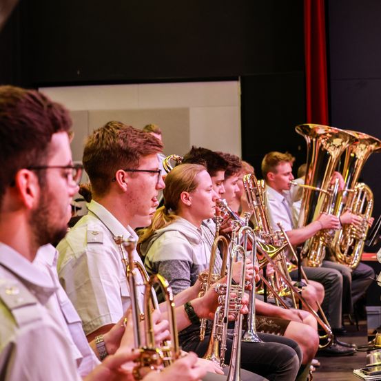 Eine Gruppe junger Musiker in weißen Uniformen spielt auf einer Bühne ihre Instrumente. Sie sitzen und stehen, alle konzentrieren sich auf ihre Instrumente. Dazu gehören Trompeten, Posaunen und Tuben.