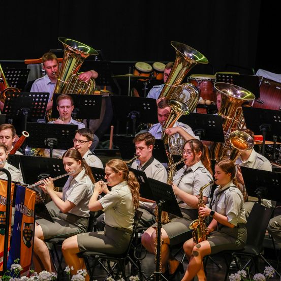 Ein Orchester spielt auf der Bühne mit verschiedenen Instrumenten wie Trompeten, Tubas und Flöten. Die Orchestermitglieder tragen Uniformen und sitzen auf Stühlen. Sie spielen vor einem dunklen Hintergrund.