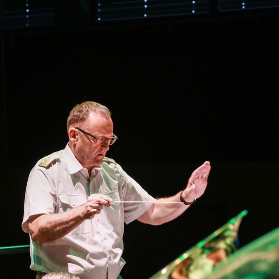 Ein älterer Mann in einer Militäruniform steht auf der Bühne, hält einen Taktstock und scheint ein Orchester zu dirigieren.