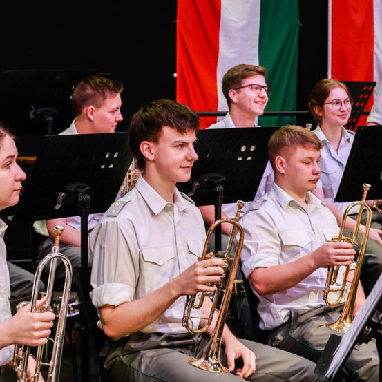 Eine Gruppe junger Musiker in Uniformen, einschließlich Trompeten, sitzt in einer Reihe auf der Bühne, mit drei Flaggen dahinter. Sie alle schauen nach vorne, wahrscheinlich in Vorbereitung auf eine Aufführung.