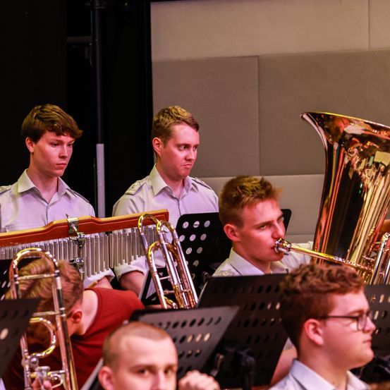 Eine Band spielt mit Blechblasinstrumenten. Ein Mann spielt Tuba, während andere Trompeten und Posaunen spielen. Sie befinden sich in einem Raum mit grauer Wand.