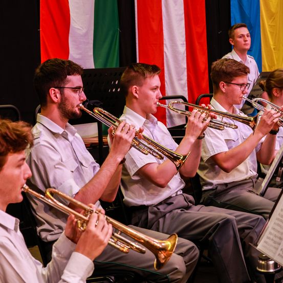 Fünf junge Männer in weißen Uniformen spielen Trompeten, während sie sitzen. Dahinter hängen Flaggen in verschiedenen Farben und Designs.
