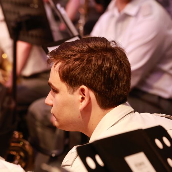 Ein Mann in einem weißen Uniform sitzt in einem Orchester und sieht konzentriert aus. Dahinter spielen verschwommene Musiker Trompeten und Posaunen.