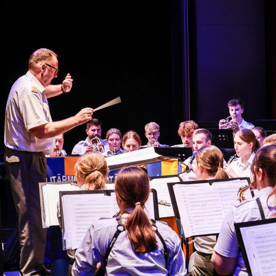 Ein Dirigent führt eine Marschkapelle mit Trompeten, Posaunen und Klarinetten. Die Musiker konzentrieren sich auf ihre Notenblätter, während sie spielen.