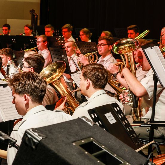 Eine Brass-Band spielt auf der Bühne, wobei die Mitglieder Blechblasinstrumente und Notenständer halten. Sie tragen Uniformen.