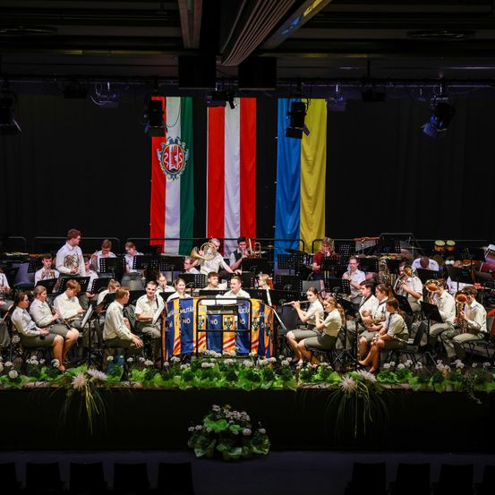 Ein Militärorchester tritt auf der Bühne auf, mit Flaggen verschiedener Nationen im Hintergrund. Die Musiker sitzen in Stühlen und spielen eine Vielzahl von Instrumenten. Die Bühne ist mit Grünpflanzen und Blumen geschmückt.