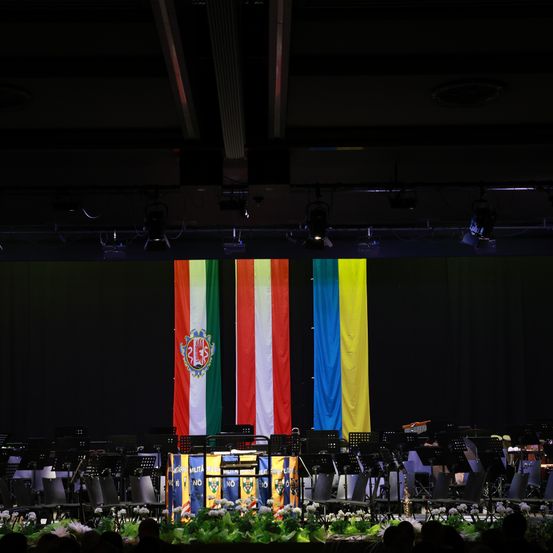 Ein Orchester auf der Bühne mit großen Fahnen Italiens und der Ukraine im Hintergrund. Die Bühne ist dunkel, mit Lichtern, die auf die Instrumente und Fahnen gerichtet sind.