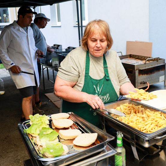 Eine Frau in einem Schürze serviert Essen bei einer Outdoor-Veranstaltung. Vor ihr stehen Tabletts mit Sandwiches und Pommes. Zwei Männer hinter ihr bereiten Essen zu.