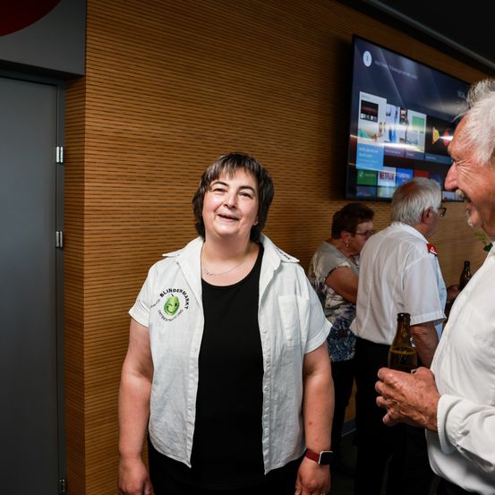 Eine lächelnde Frau steht in der Nähe einer Wand mit einem Fernsehmonitor im Hintergrund, hält eine Bierflasche und unterhält sich mit einer anderen Person.