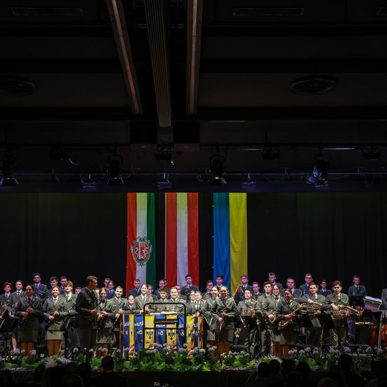Eine Gruppe von Musikern in Uniform, einschließlich eines Dirigenten, tritt mit Flaggen und Instrumenten auf der Bühne auf. Das Publikum beobachtet sie.