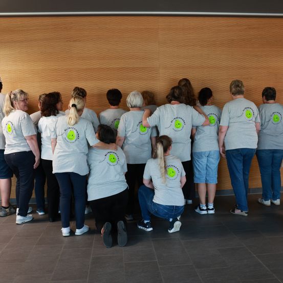 Eine Gruppe von Frauen in grauen T-Shirts mit lächelnden Gesichtern, die in einer Reihe stehen und sitzen und sich einem hölzernen Hintergrund zuwenden. Sie nehmen wahrscheinlich an einer Veranstaltung oder einem Treffen teil.