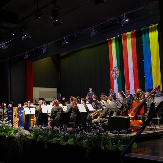 Ein Militärorchester spielt auf einer Bühne vor einem Flaggenhintergrund. Musiker in Uniform spielen verschiedene Instrumente, darunter Trompeten, Celli und Trompeten. Die Bühne ist mit Blumen geschmückt, und Scheinwerfer sind an der Decke montiert.