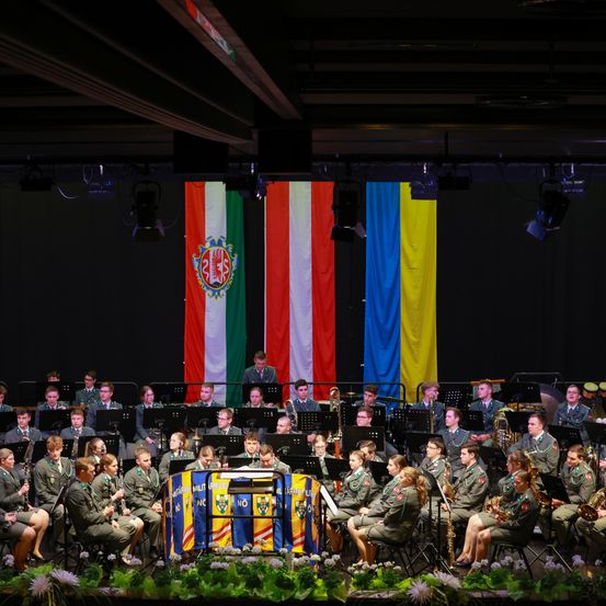 Ein Militärorchester spielt auf der Bühne. Im Hintergrund befinden sich Flaggen aus Ungarn und der Ukraine. Die Mitglieder sitzen in Reihen und spielen verschiedene Instrumente.