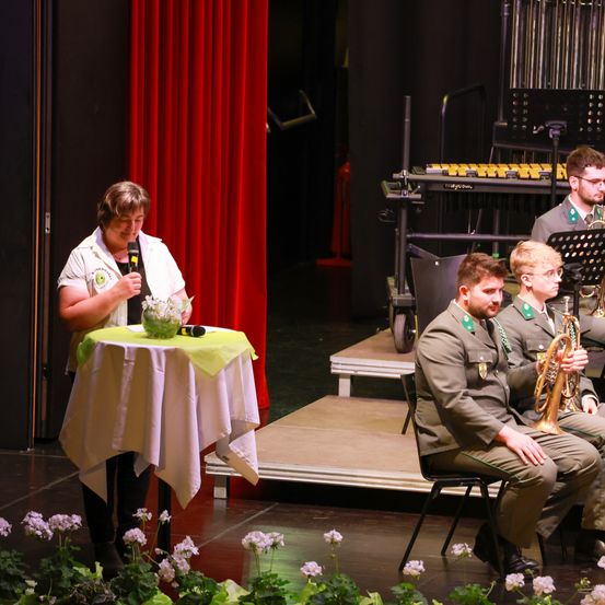Eine Frau mit einem Mikrofon steht mit einem Tisch und Blumen auf der Bühne. Dahinter ist ein roter Vorhang. Zwei Männer in Uniform sitzen auf einem Stuhl und spielen Musikinstrumente.