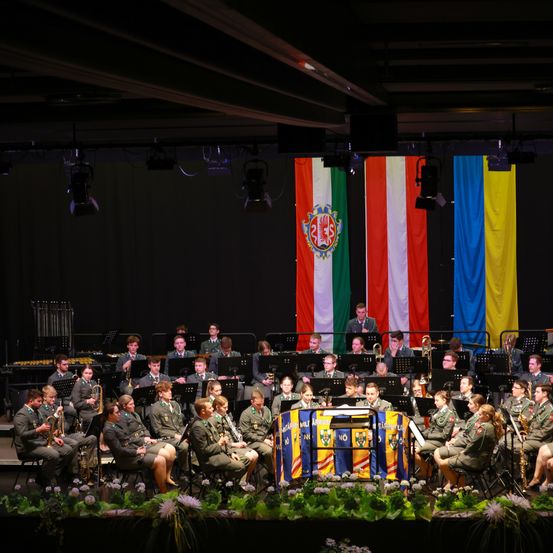 Ein Militärorchester tritt auf der Bühne auf, mit einem Hintergrund von Flaggen, eine mit einem Logo und zwei gestreiften Flaggen. Die Orchestermitglieder sind in Uniform und spielen verschiedene Instrumente, sitzend und stehend.