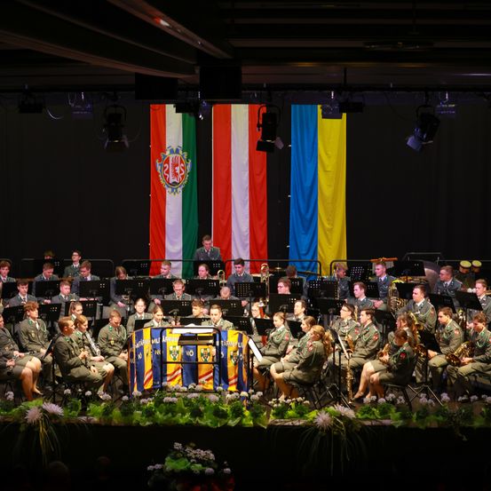 Ein Militärorchester spielt auf der Bühne mit Musikinstrumenten. Die Bühne ist mit Flaggen und Blumen geschmückt. Die Bandmitglieder sind in Uniform und sitzend. Es gibt Scheinwerfer an der Decke.