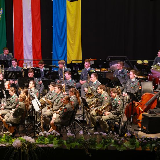 Ein Militärorchester in Uniformen spielt auf der Bühne mit verschiedenen Instrumenten wie Trompeten, Posaunen und Trommeln, mit Flaggen und Blumen als Hintergrund.