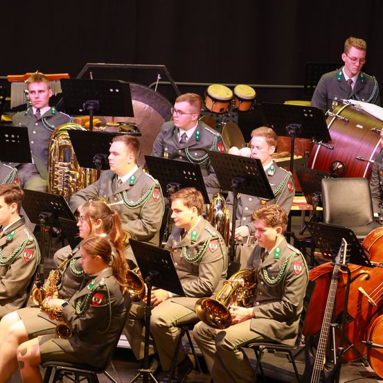 Ein Militärorchester in Uniform spielt auf der Bühne. Sie spielen verschiedene Instrumente wie Trommeln, Saxophone und Trompeten. Das Orchester sitzt auf Stühlen, während andere dahinter stehen.