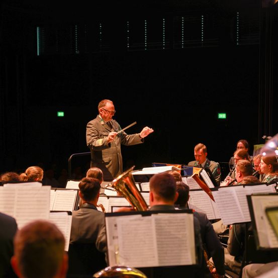 Ein Militärorchester spielt auf der Bühne, angeführt von einem Dirigenten, Musiker spielen verschiedene Instrumente und das Publikum sitzt davor.