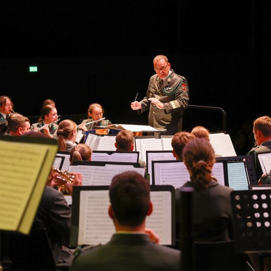 Ein Militärorchester tritt mit einem Dirigenten auf der Bühne auf. Verschiedene Instrumente werden benutzt, und das Publikum sitzt davor.