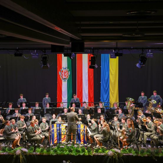 Ein Militärorchester tritt auf einer Bühne auf, im Hintergrund die Flaggen zweier Länder. Die Musiker sind in Uniform gekleidet.