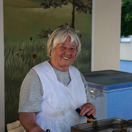 Eine Frau in einem weißen Schürze steht neben einer Edelstahl-Fritteuse und lächelt in die Kamera. Hinter ihr zeigt eine Wandmalerei ein blumiges Feld mit einem Baum.