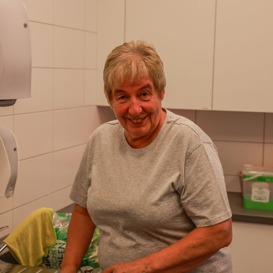 Eine Frau mit kurzen grauen Haaren steht in einer Küche und lächelt für die Kamera. Sie trägt ein graues T-Shirt und ist von Küchenutensilien umgeben.