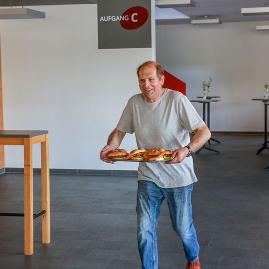 Ein Mann, der einen Tablett mit Sandwiches trägt, lächelt in einem Büro. Ein Schild an der Wand steht Aufgang C. Im Hintergrund befinden sich Tische und Stühle.