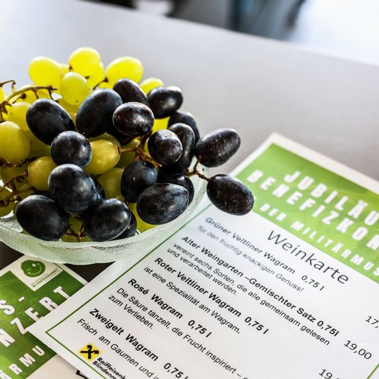 Eine Schüssel mit gemischten Trauben, einige grün und einige schwarz, steht auf einem Tisch neben einer Karte, die 'Roter Veltliner - Gemischter Wagram' lautet. Die Karte enthält Informationen über eine Weinverkostung.