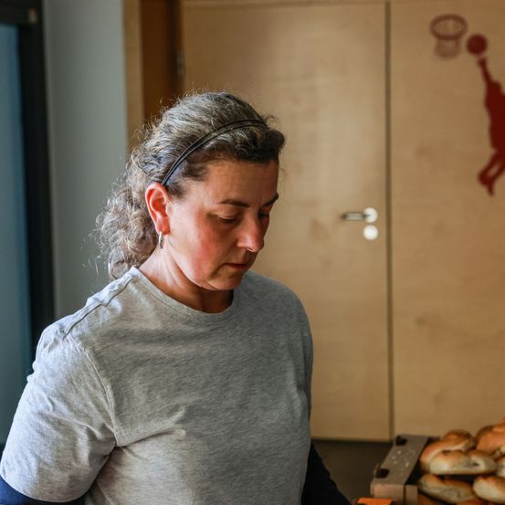 Eine Frau in einem grauen T-Shirt steht in einer Küche und blickt auf einen Tablett mit Essen. Hinter ihr ist eine Holztür mit einer Basketballsilhouette.