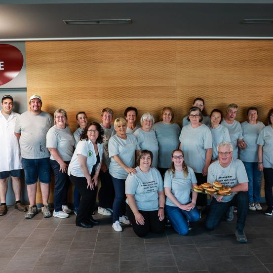 Eine Gruppe von Menschen in grauen T-Shirts posiert für ein Foto. Einige halten Essen. Hinter ihnen ist eine Holzwand zu sehen.