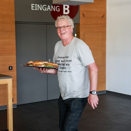 Ein lächelnder Mann mit Brille und Uhr hält einen Tablett mit Sandwiches und steht in einem Flur mit einer Holzwand und einem Schild, das 'EINGANG B' steht.