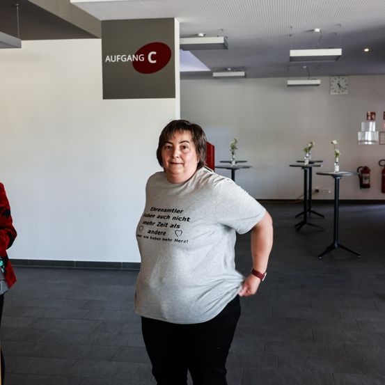 Eine Frau steht in einem Raum mit weißen Wänden und leerem Innenraum, trägt ein graues T-Shirt mit gedrucktem Text. Hinter ihr steht ein Schild mit der Aufschrift 'AUFGANG C'. Tische und Stühle sind im Raum aufgestellt.