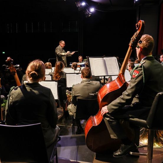 Ein Militärorchester spielt auf der Bühne, angeführt von einem Dirigenten, Musiker spielen verschiedene Instrumente, und Zuschauer sehen vom Publikum aus zu.