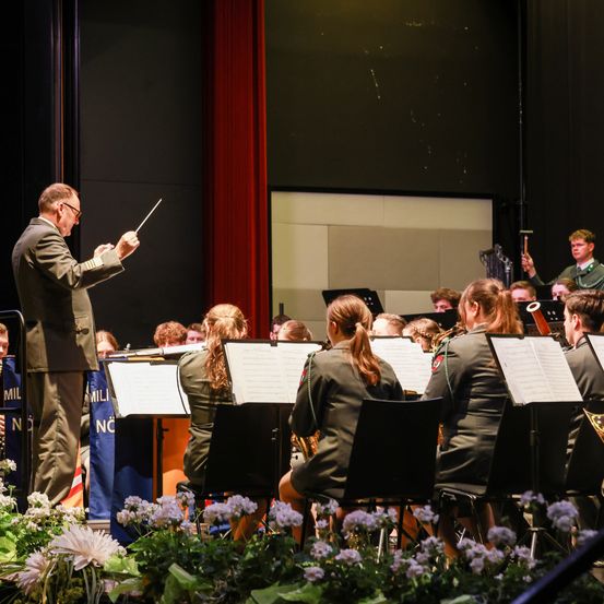 Ein Orchester spielt auf einer Bühne, angeführt von einem Mann in Uniform. Die Musiker sitzen auf Stühlen und spielen Instrumente. Die Bühne hat einen roten Vorhang, einen Pult und eine Blumendekoration.