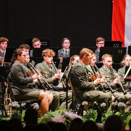 Ein Militärorchester spielt auf der Bühne, mit Musikern, die Klarinetten, Trompeten und andere Instrumente spielen, vor einer Flagge.