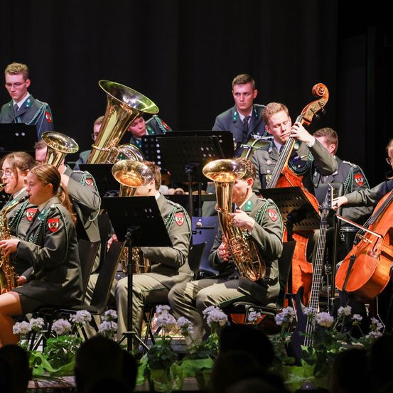 Eine Gruppe von Militärmusikern spielt verschiedene Instrumente, darunter Tuben, Celli und Saxophone, auf der Bühne. Das Publikum sitzt vor ihnen und verfolgt die Aufführung.