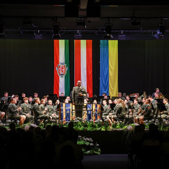 Ein Militärorchester spielt auf der Bühne, mit einer Flagge im Hintergrund. Das Publikum sitzt davor und beobachtet die Aufführung.