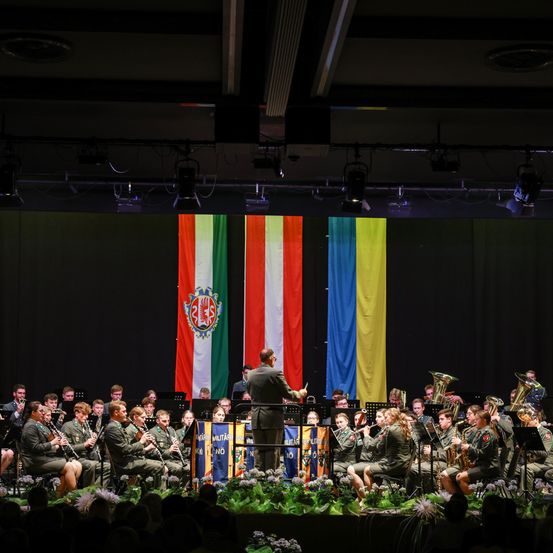 Ein Militärorchester spielt auf einer Bühne mit Flaggen im Hintergrund. Die Band besteht aus Klarinetten, Trompeten und Tuben. Ein Dirigent steht vor der Band.