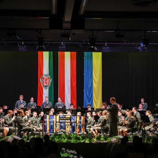 Ein Militärorchester spielt auf der Bühne, mit den Flaggen von Österreich und der Ukraine im Hintergrund. Der Dirigent dirigiert die Musiker, während sie Trompeten, Flöten und andere Instrumente spielen.