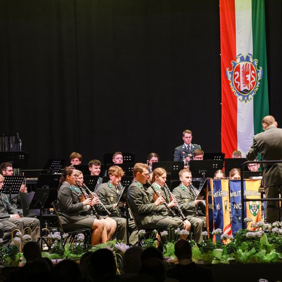 Ein Militärorchester spielt auf der Bühne, Musiker spielen Klarinetten und Trompeten. Ein Publikum beobachtet von vorne. Eine Flagge und ein Banner sind hinter der Band angebracht.