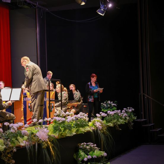 Eine Bühnenaufführung mit einem Mann in Uniform, einer Frau, die von einem Skript liest, und einer Band mit Musikinstrumenten. Die Bühne ist mit Blumen und einem roten Vorhang geschmückt.