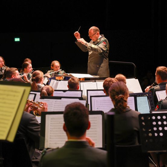 Ein Militärorchester spielt auf der Bühne mit einem Dirigenten. Die Musiker sind in Uniform gekleidet und spielen verschiedene Instrumente.