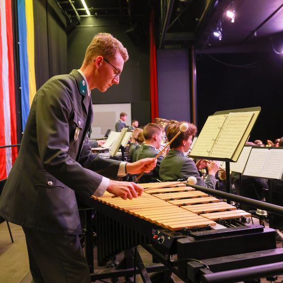 Ein Mann in einer Militäruniform spielt Xylophon in einem Musikraum mit anderen Musikern und einer Flagge.