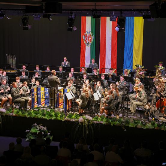 Ein Militärorchester spielt auf der Bühne vor Flaggen im Hintergrund, unter Scheinwerfern. Musiker spielen verschiedene Instrumente, einige sitzend und andere stehend. Das Publikum sitzt davor.