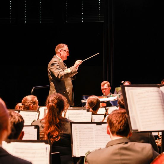 Ein Orchesterdirigent in Uniform führt ein Blasorchester auf der Bühne an, mit Musikern vor ihm sitzend.