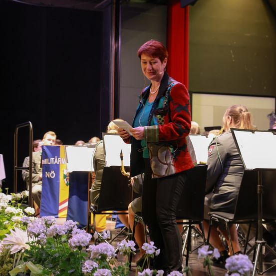 Eine Frau steht auf der Bühne und hält ein Blatt Papier in der Hand. Hinter ihr befindet sich ein Militärorchester. Ein blauer Fahnenmast mit weißem Text steht hinter den sitzenden Musikern.