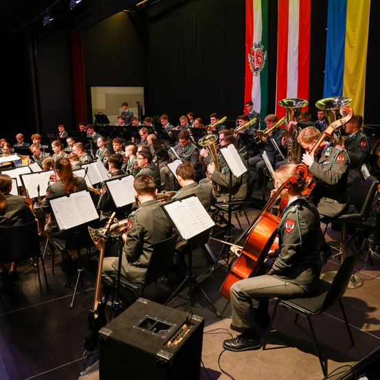 Ein Militärorchester probet auf der Bühne mit Fahnen im Hintergrund. Die Mitglieder sitzen und spielen verschiedene Instrumente.