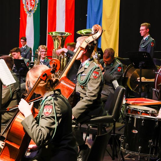 Ein Militärorchester tritt auf der Bühne auf. Die Mitglieder spielen Instrumente wie Cello, Tuba und Trommeln. Sie tragen Uniformen mit Fahnen im Hintergrund.