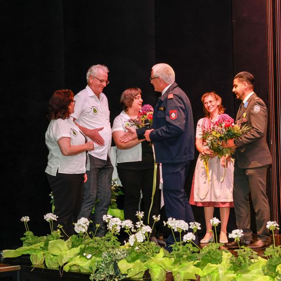 Eine Gruppe von Menschen steht mit Blumen auf der Bühne. Zwei Männer in Uniformen halten Blumen, während die anderen zusehen. Eine Frau hält ein Band.